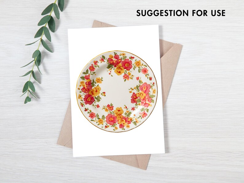 Pode incluir: Um prato de cer&acirc;mica branca com borda dourada, adornado com um padr&atilde;o floral em tons de vermelho, laranja e amarelo. O prato &eacute; apresentado em uma superf&iacute;cie branca com um envelope marrom e um galho de vegeta&ccedil;&atilde;o.