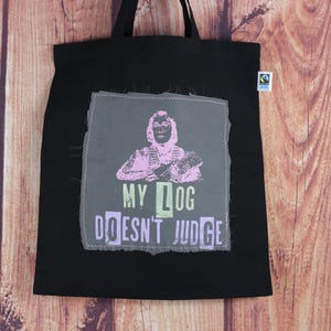 Könnte beinhalten: Schwarze Tragetasche mit einem grafischen Aufnäher. Der Aufnäher zeigt eine rosa und lila Illustration einer Person und den Text "MY LOG DOESN'T JUDGE" in Grün und Lila. Die Tasche hat schwarze Griffe und ein kleines Etikett.