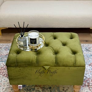 May include: An olive green velvet ottoman with button detailing and wooden legs. A gold-coloured tray with a candle and diffuser sits on top. The text "Fluffy Pouffe" is on the front.