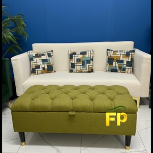 May include: A cream-colored sofa with three decorative pillows featuring geometric patterns. In front of the sofa is a green tufted ottoman with black legs and gold accents. The background is a solid blue wall.