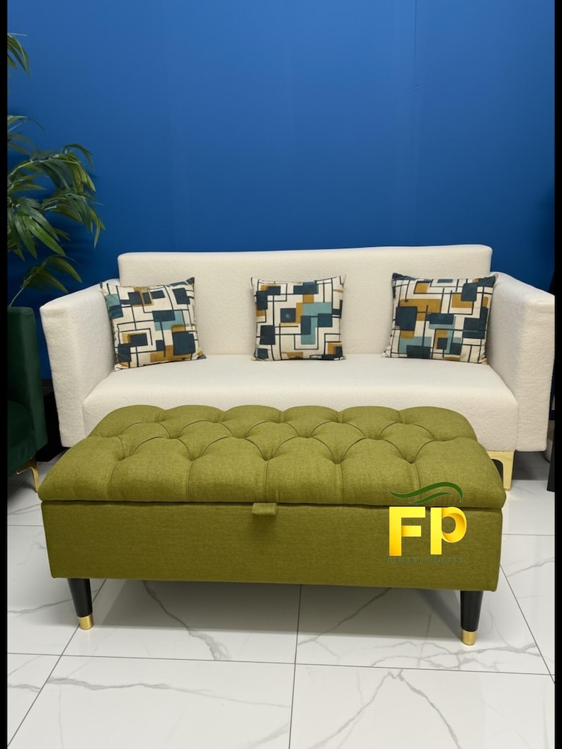May include: A cream-colored sofa with three decorative pillows featuring geometric patterns. In front of the sofa is a green tufted ottoman with black legs and gold accents. The background is a solid blue wall.