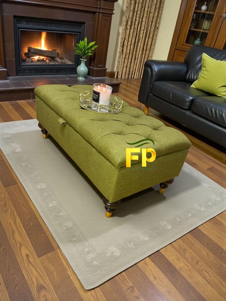 May include: A tufted, olive green storage ottoman with dark wooden legs and gold accents. The rectangular ottoman sits on a light beige rug, with a lit fireplace in the background. A candle sits on a decorative tray on top of the ottoman.