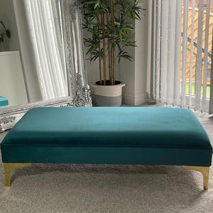May include: A teal velvet ottoman with gold metal legs. The ottoman is sitting on a grey carpet.