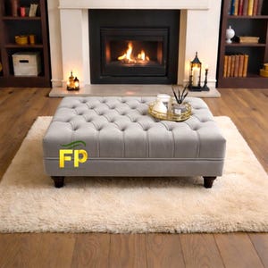 May include: A grey, buttoned ottoman with dark wooden legs on a cream rug. A gold tray with candles and a diffuser sits on the ottoman. The ottoman is in front of a fireplace with a burning fire.