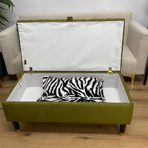 May include: An olive green storage ottoman with a white interior and a zebra-print pillow. The ottoman has dark wooden legs with gold tips and a hinged lid. A cream-colored sofa is in the background.
