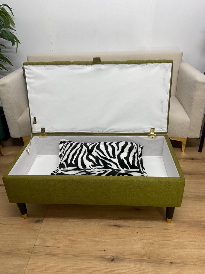 May include: An olive green storage ottoman with a white interior and a zebra-print pillow. The ottoman has dark wooden legs with gold tips and a hinged lid. A cream-colored sofa is in the background.