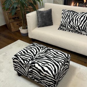 May include: A zebra-print ottoman sits on a white rug, with a cream-coloured sofa behind it. A cushion with the same zebra pattern and a grey cushion are on the sofa. A fireplace and a potted plant are in the background.