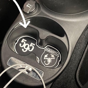 Può includere: Sottobicchieri neri per portabicchieri per auto con testo bianco "595" e logo Abarth. L'immagine mostra anche una presa di corrente da 12 V CC e una porta di ricarica USB.