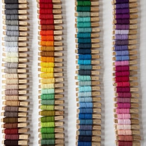 May include: A collection of 300 embroidery floss spools organised by colour. The spools are arranged in rows on a white background. Each spool is labelled with the colour name. The colours include black, grey, white, red, orange, yellow, green, blue, purple, pink, and brown.