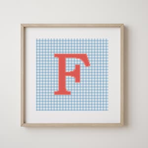 May include: Framed artwork with a cross-stitch design of the letter "F" in coral. The letter is set against a light blue and white gingham pattern. The artwork is in a light-coloured wooden frame, suitable for a nursery or a child's room.