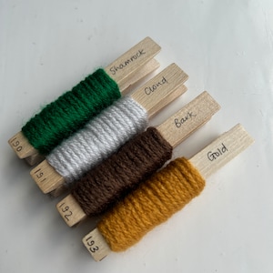 May include: Five spools of yarn on wooden pegs labelled with the colours "Shamrock", "Cloud", "Bark", "Gold", and corresponding numbers 190, 191, 192, and 193.