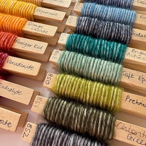 May include: Close-up of yarn wound around wooden spools labelled with mineral names, including Stilbite, Aragonite, Orpiment, Orange Calcite, Realgar Red, Vanadinite, Endralyte, Lazurite, Rosy, Sodalite, Bright Actinolite, Soft Epidote, Prehnite, Chlorite Green, and Granite.