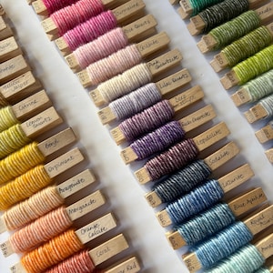May include: A collection of yarn skeins wrapped around wooden spools, each labelled with a different gemstone name. The yarn colours range from deep red to light blue, with a variety of shades in between.