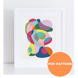 May include: A colorful abstract cross stitch pattern with a variety of shapes and colors, including pink, blue, green, yellow, and orange. The pattern is framed in a white frame. The text "PDF PATTERN" is on a bright orange circle.