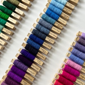 May include: A collection of yarn samples in various colors, including greens, blues, and purples, are displayed on wooden sticks. Each stick has a small label with the yarn color name. The arrangement is on a white surface, showcasing the range of hues.