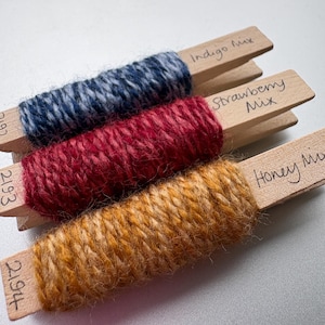 May include: Three wooden pegs, each wrapped with a different colour of yarn. The yarn colours are indigo, red, and golden yellow. Each peg has handwritten text indicating the yarn colour mix: "Indigo Mix", "Strawberry Mix", and "Honey Mix".