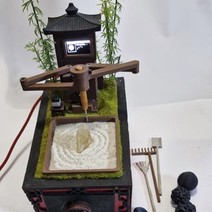 May include: A miniature zen garden with a wooden pagoda, bamboo stalks, and a sand tray with a rock. The garden includes small rakes, decorative stones, and a small screen. The base is black with red accents.