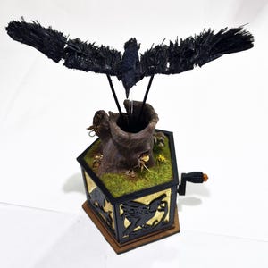 May include: A handcrafted music box with a black raven sculpture above a tree trunk. The box has a gold and black design with dragon and raven silhouettes. The base is brown, and the top has a green, mossy appearance.