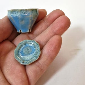 Può includere: Un set in miniatura composto da tazzina e piattino in ceramica con forma esagonale sfaccettata. La ceramica presenta uno smalto reattivo lucido nei toni dell'azzurro cielo e del verde acqua, con una texture maculata e base in argilla chiara.