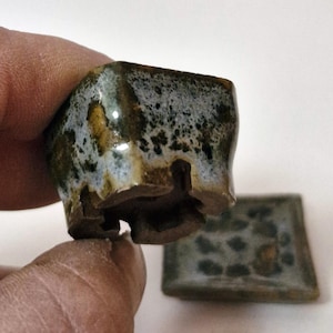 May include: A small square ceramic bonsai pot with a mottled glaze in shades of blue, green, and brown. The pot features a thick rim, a textured exterior surface, and a base with four feet and drainage openings. A matching square tray is included.