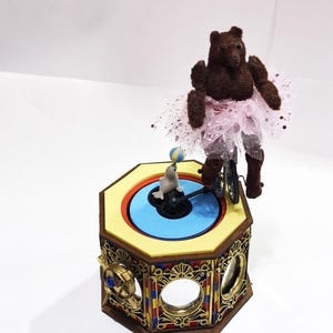 May include: A whimsical music box featuring a brown bear in a pink tutu riding a miniature bicycle. A seal balances a ball on its nose. The box is octagonal with ornate gold and colorful accents, creating a circus theme.