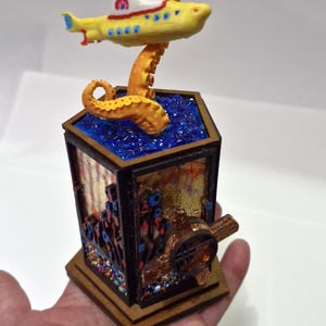 May include: A whimsical, handcrafted art piece featuring a yellow submarine with a red and white top, perched above a blue, textured base. An orange octopus tentacle supports the submarine. The base is a wooden box with colorful embellishments.