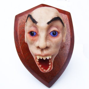 Könnte beinhalten: Ein groteskes, blasses Gesicht mit blauen Augen und einem breiten, zahnigen Grinsen ist auf einer dunklen Holzplatte montiert. Das Gesicht ist realistisch gestaltet, mit sichtbaren Poren und Falten.