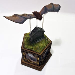 May include: A miniature diorama featuring a bat with outstretched wings, mounted above a tombstone on a hexagonal base. The base is decorated with faux grass and framed landscape images, creating a spooky, gothic theme.