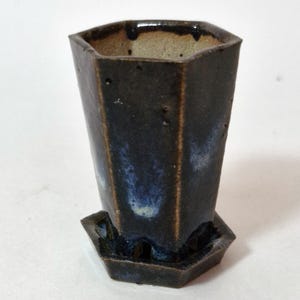 May include: A hexagonal ceramic planter with a dark brown and blue glazed finish, featuring a matching hexagonal saucer base. The interior of the pot is a light beige colour, and the exterior surface has a textured, rustic appearance.