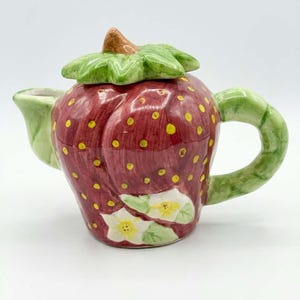 Può includere: Una teiera in ceramica a forma di fragola con finitura rosso lucido, semi gialli e coperchio a foglie verdi. Il manico e il beccuccio sono verdi, e la base presenta fiori di fragola bianchi dipinti con centri gialli su sfondo rosso.