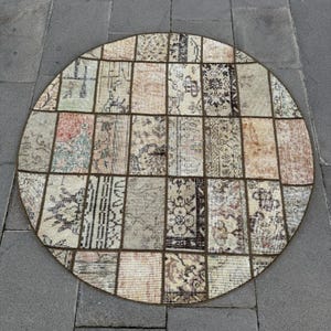 Puede incluir: Alfombra redonda de patchwork con una variedad de colores apagados, incluyendo beige, marrón y rojos descoloridos. La alfombra está hecha de piezas rectangulares y cuadradas individuales, creando un patrón geométrico. Se encuentra sobre una superficie de piedra gris.