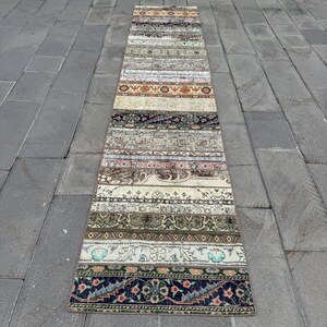Peut inclure: Un long tapis de couloir étroit présentant un motif en patchwork composé de multiples bandes horizontales. Le tapis affiche une variété de motifs et de couleurs complexes, notamment des tons de bleu, orange, vert, crème et brun, avec des motifs floraux et géométriques.