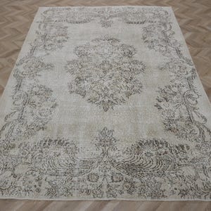 Peut inclure: Un tapis rectangulaire au fond beige clair orné d'un motif complexe de fleurs et de vignes brun foncé. Le design présente un grand médaillon central entouré d'une bordure détaillée composée de feuillages et de motifs botaniques.