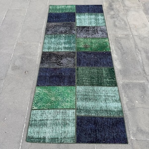 May include: A rectangular patchwork rug with a variety of green and blue hues. The rug is composed of rectangular patches of varying shades, from deep navy to light teal. The rug is placed on a gray stone surface.