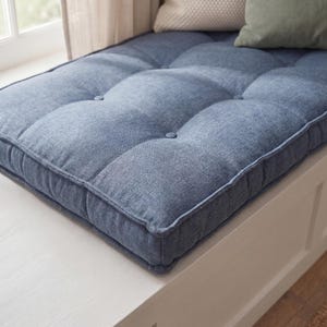 May include: A rectangular blue tufted bench cushion with a textured fabric finish. The cushion features a grid of fabric-covered buttons across its surface, creating a quilted appearance. It is positioned on a white wooden base with a flat edge.