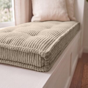 May include: A rectangular beige corduroy bench cushion featuring a tufted design with visible vertical ridges. The cushion has thick, piped edges and a soft, textured fabric surface, providing a structured appearance for a seating area.