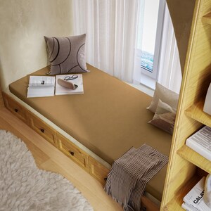 May include: A cozy, built-in bed with a tan mattress, a decorative pillow, and a striped blanket. The bed has wooden drawers and is next to a window with white curtains. Books and glasses rest on the bed.