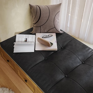 May include: A black leather bench with tufted detailing, a light brown pillow with a dark brown circular pattern, a book, glasses, and a glasses case. The bench is placed near a window with white curtains.