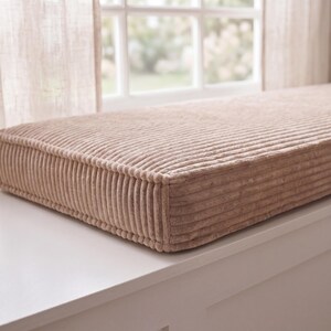 Puede incluir: Un cojín rectangular para banco de pana en color marrón claro. El cojín tiene una textura acanalada y está colocado sobre una superficie blanca cerca de una ventana con cortinas transparentes. El cojín mide aproximadamente 76 cm de largo.