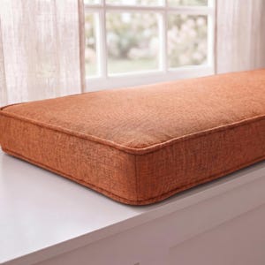 May include: A rectangular bench cushion upholstered in a textured, burnt orange fabric. The cushion features piped edges along the top and bottom perimeter, creating a structured, boxy shape that sits flat on a smooth, white surface.