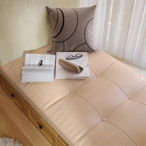May include: A light beige tufted bench with integrated wooden drawers, topped with a square gray pillow featuring a dark circular pattern, an open book, a closed book, a pair of black-rimmed glasses, and a beige hard-shell glasses case.