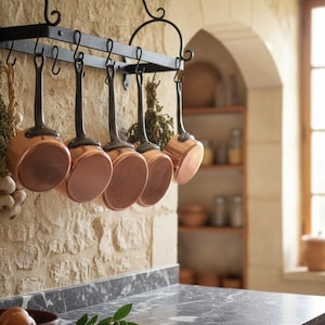 Puede incluir: Escena de cocina con un colgador de ollas de hierro forjado negro con seis cacerolas de cobre. Se ven manojos de hierbas y ajo. Las cacerolas tienen mangos oscuros y están colgadas contra una pared de piedra texturizada.