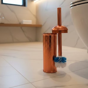Puede incluir: Un juego de cepillo y soporte para inodoro de color cobre. El soporte tiene una textura martillada, y el cepillo presenta cerdas azules y blancas. El conjunto se exhibe en un baño moderno.