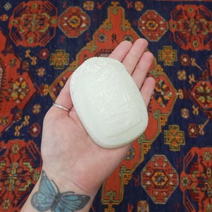 May include: A white, rectangular, carved stone with rounded edges. The stone features detailed relief carvings of figures and symbols. The object is held in a hand, with a blue butterfly tattoo visible on the wrist.