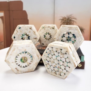 May include: Five hexagonal boxes made of white mother of pearl with intricate geometric patterns in green and blue. The boxes are arranged in a semi-circle on a white surface.