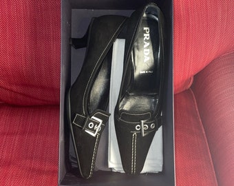 Könnte beinhalten: Ein Paar schwarze Wildlederschuhe mit Absatz, Schnallenverzierung und weißen Nähten. Die Schuhe befinden sich in einer Schachtel mit dem Prada-Logo und dem Text "Made in Italy".