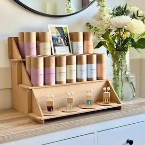 May include: A tiered wooden display unit showcasing car air fresheners in cylindrical containers with pastel labels. Small glass bottles with wooden toppers and cork bases are also displayed. A framed picture and a vase of flowers are visible in the background.