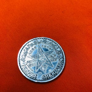 May include: A silver coin with a five-pointed star in the center. The star has a crown in the center and the words "EMPIRE CHERIFIEN" around the outside. The coin also has the years "1956" and "1376" on it.