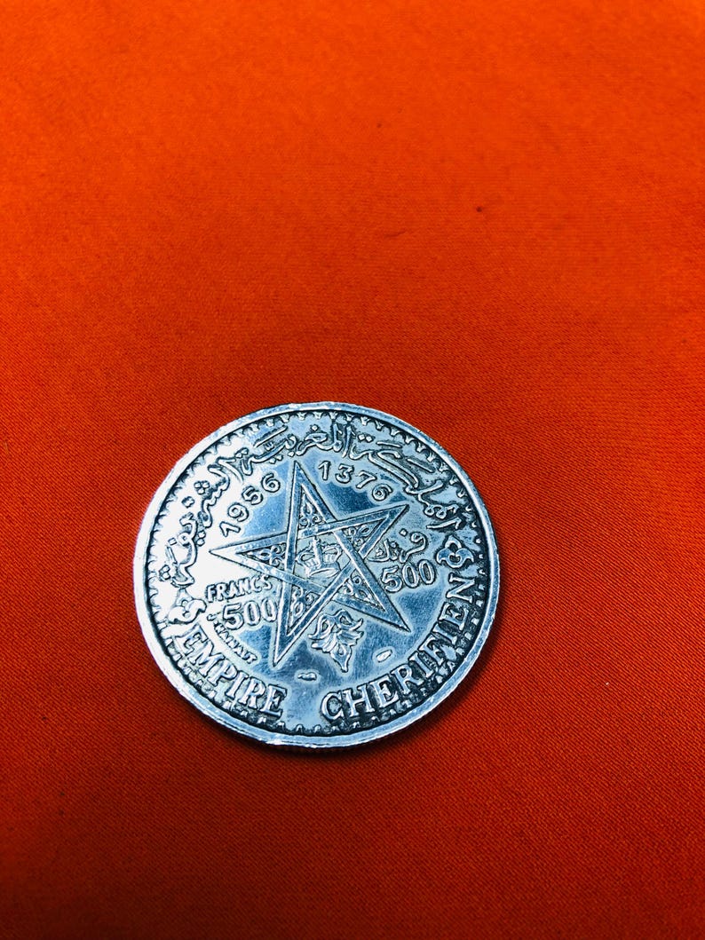 May include: A silver coin with a five-pointed star in the center. The star has a crown in the center and the words "EMPIRE CHERIFIEN" around the outside. The coin also has the years "1956" and "1376" on it.