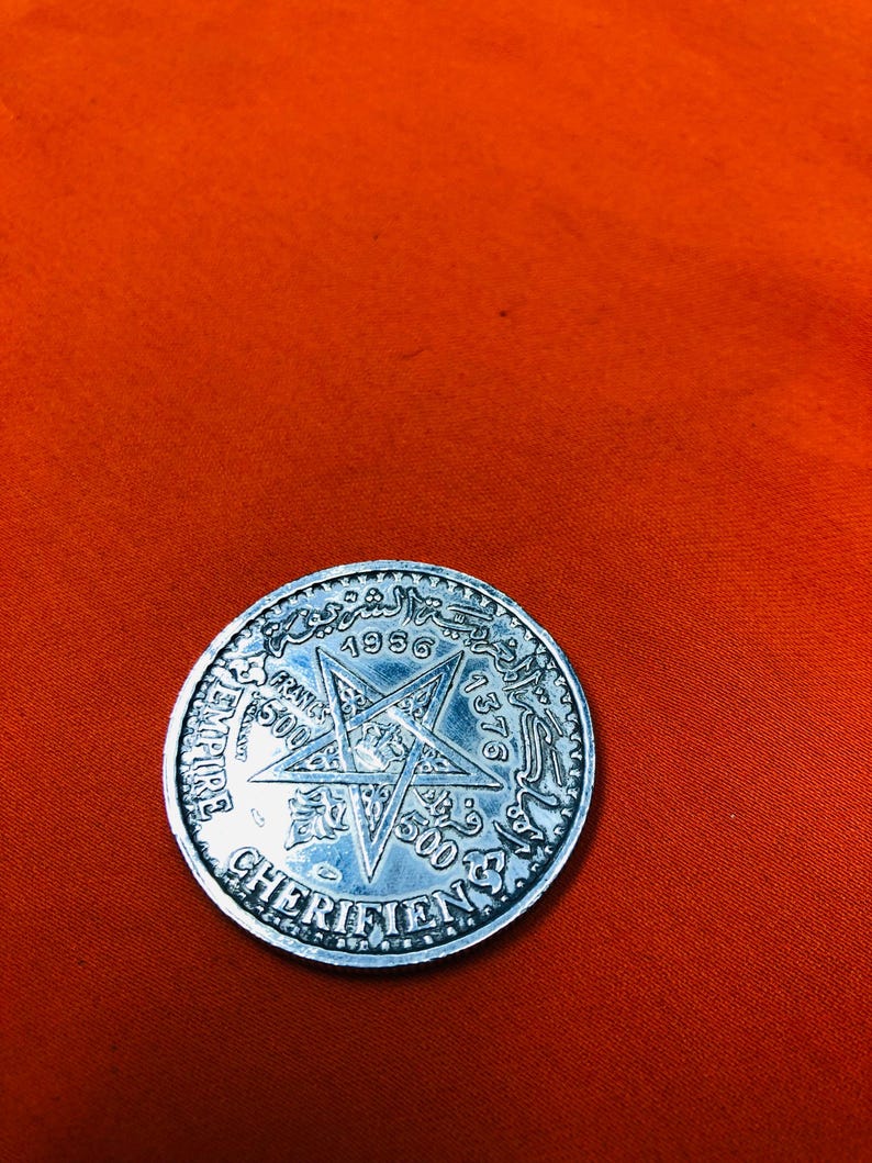 May include: A silver coin with a five-pointed star in the center and the text "1376 1956" around the star. The text "EMPIRE CHERIFIEN" is around the edge of the coin.
